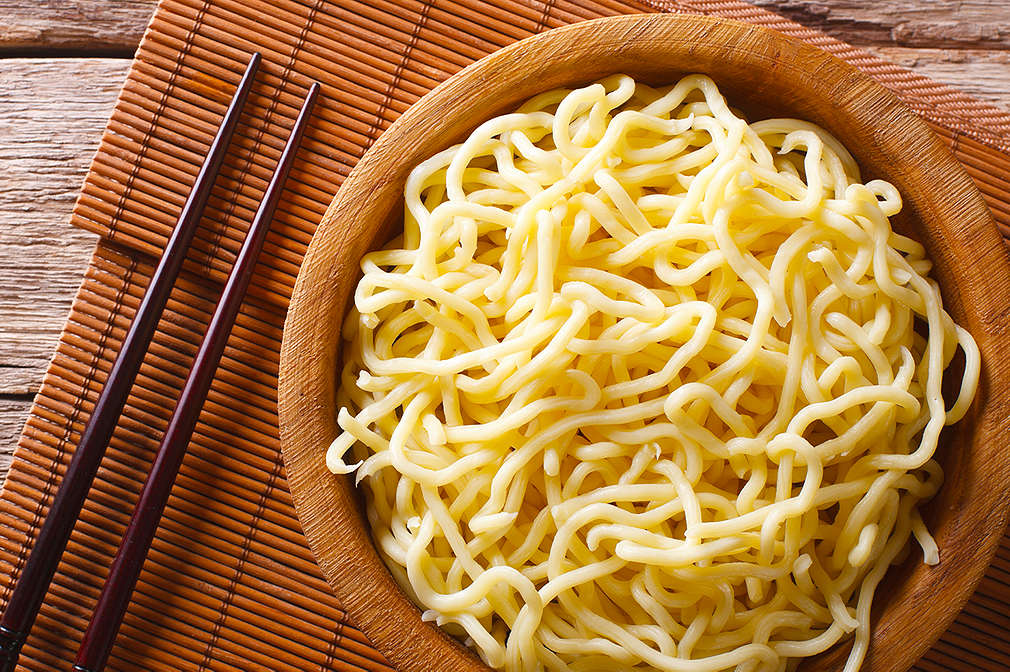 Dunkler Holztisch, auf dem ein braunes Platzdeckchen aus Bambus liegt. Darauf liegen ein Paar Stäbchen und eine Holzschale gefüllt mit gekochten Ramen-Nudeln