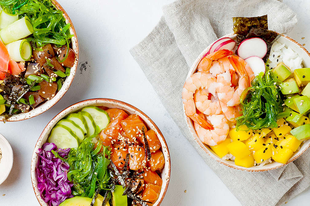 Drei Bowls mit unterschiedlichen Inhalten, unter anderem Thunfisch, Garnelen, Avocado, Gurke, rote Zwiebeln, Radieschen, Algen. Außerdem auf dem Tisch: graues Geschirrtuch, Schälchen mit Sesam, Kimchi und Seetang