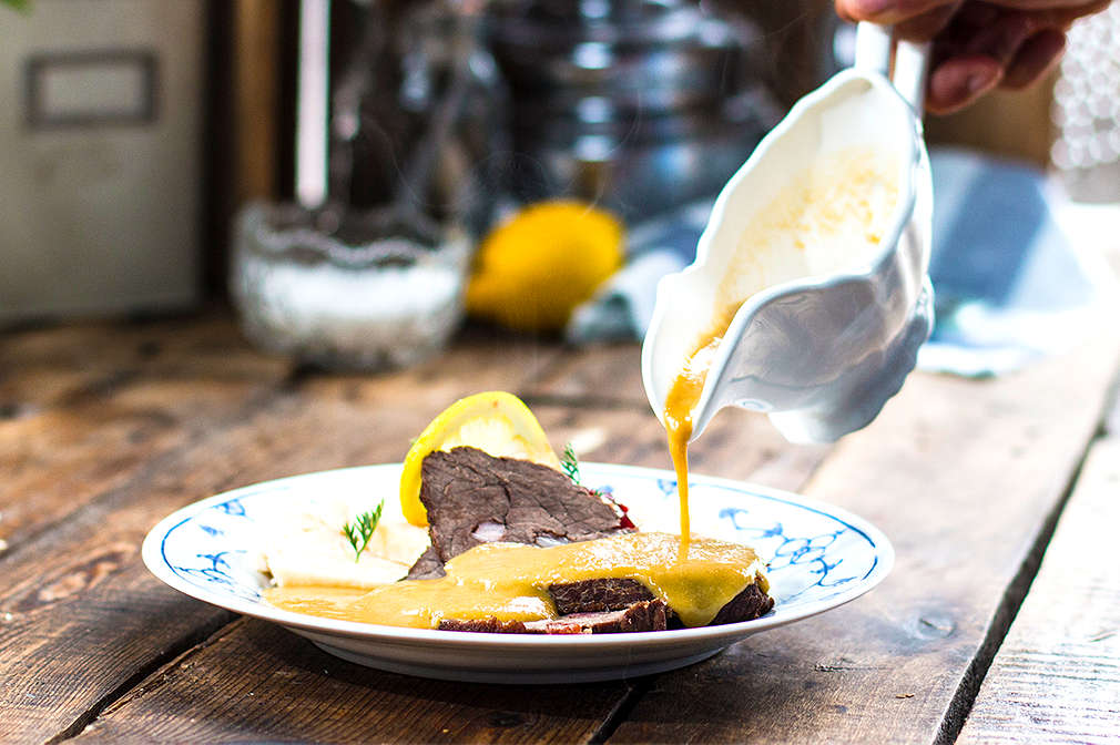 Eine Hand gießt Soße über eine Portion Svíčková mit Böhmischen Knödeln, die sich auf einem Teller befindet, der auf einem Holztisch steht