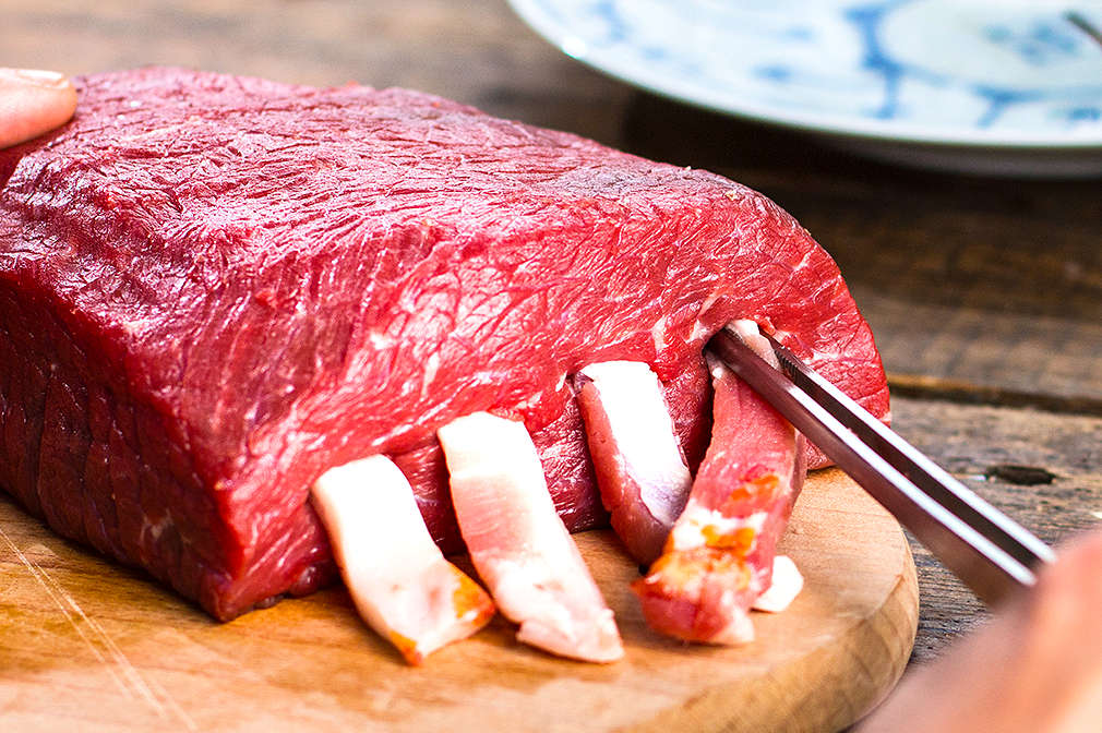 Eine Hand steckt mithilfe einer Pinzette Speckstränge in den durchstochenen Rinderbraten, der auf einem Holzbrett liegt