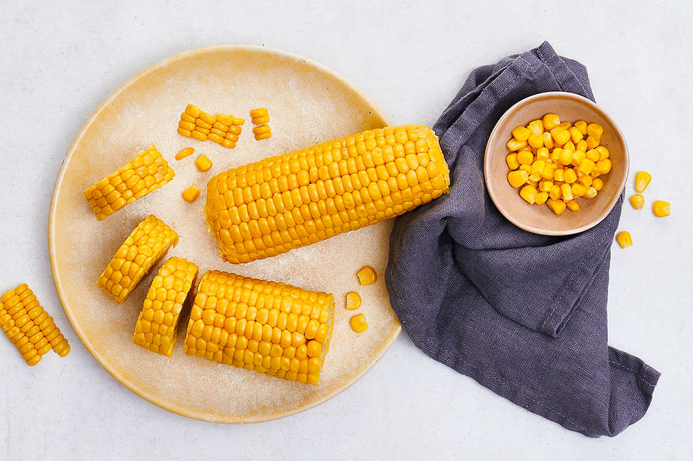 Frischer Zuckermais am Kolben, in Scheiben und in einzelnen Körnern auf einem hellen Teller mit schwarzer Serviette daneben
