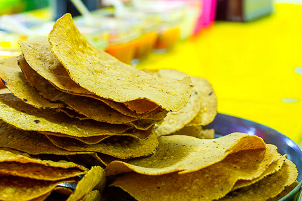 Zu sehen sind einige Maistortillas auf einem Teller, der Teller steht auf einem gelben Tisch, der vor einem bunten Hintergrund steht