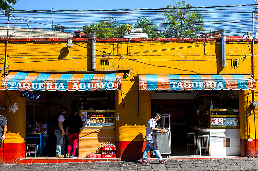 Zu sehen ist eine mexikanische Taquería mit gelber Fassade und einer blau-orange gestreiften Markise