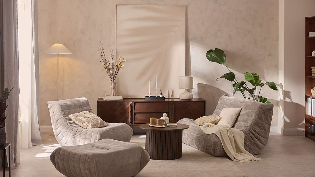 Wohnzimmer mit heller Einrichtung und diversen Möbeln wie zwei Sessel, einem Hocker, einem runden Wohnzimmertisch und einen Sideboard. An der Wand hängt ein Bild mit einer Blume.