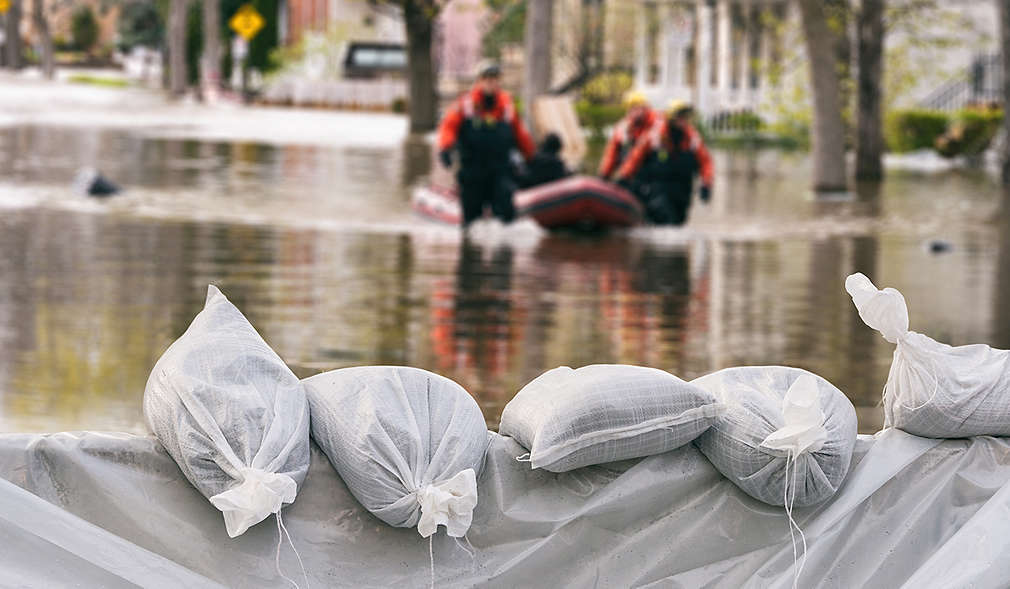 Staumauer aus Säcken gegen Hochwasser, im Hintergrund Rettungskräfte, die eine Person aus dem Hochwasser bergen