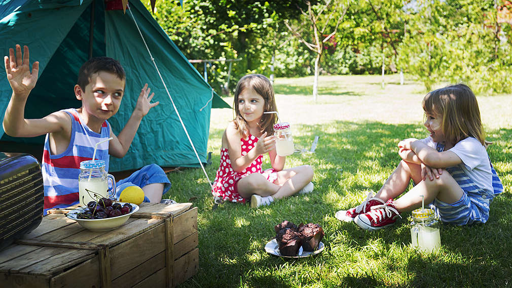 Un băiat și două fete stau în fața unui cort pe pajiște, vorbesc și beau limonadă. În fața cortului este un suport de lemn pe care se află un pahar de limonadă, o lămâie, un bol de cireșe și un radio.