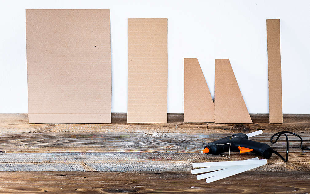 Na rustikálním dřevěném podkladu jsou kusy lepenky různých formátů opřené o bílou stěnu. Na podkladu leží tavná pistole a skládací metr.