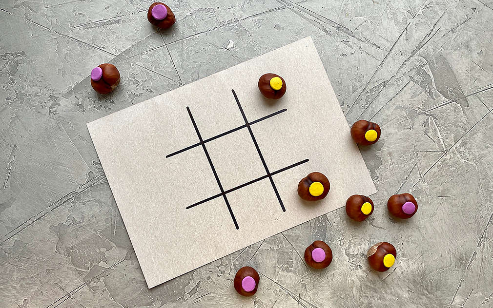 Na obrázku je: sivý štruktúrovaný podklad, na ňom biely tvrdý papier položený na šírku, na ktorom je mriežka na piškvorky. Okolo ležia hracie figúrky vytvorené z gaštanov, ktoré sú označené dvomi rôznymi farbami.
