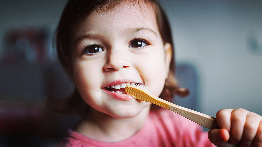 O fetiță zâmbește și se spală pe dinți cu o periuță de dinți din lemn.
