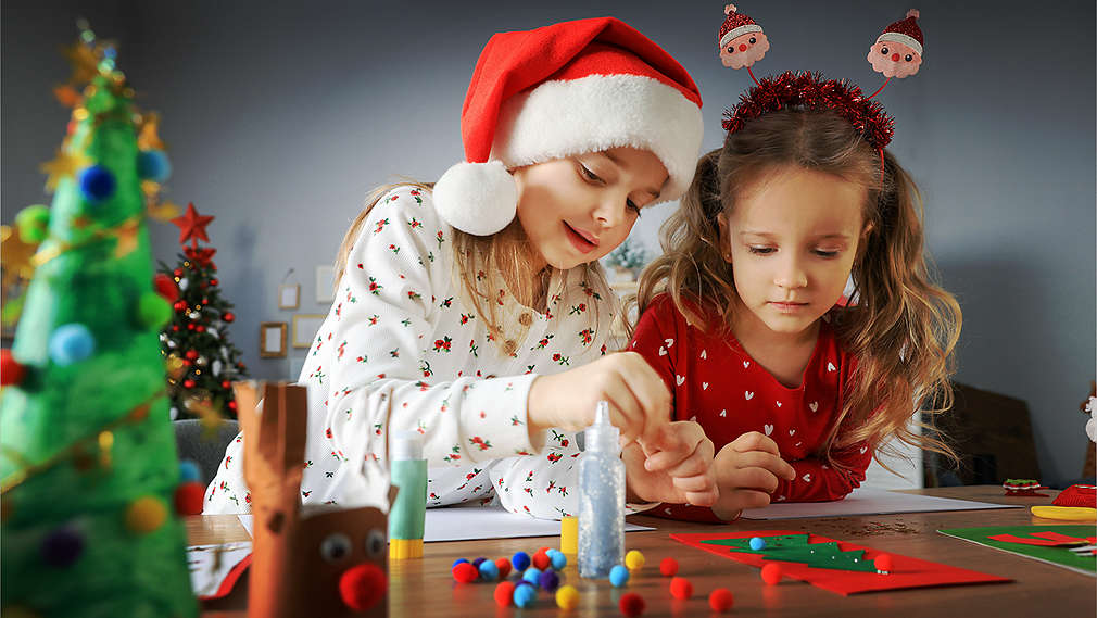 Două fetițe îmbrăcate de Crăciun stau la o masă decorată și confecționează felicitări.