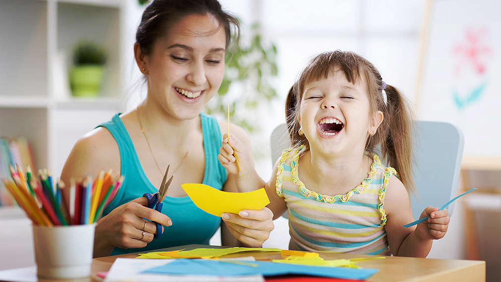 O mamă și fiica ei stau și bricolează la o masă pe care se află o cană cu creioane colorate și hârtie colorată în diferite culori. Mama taie o bucată de hârtie galbenă.