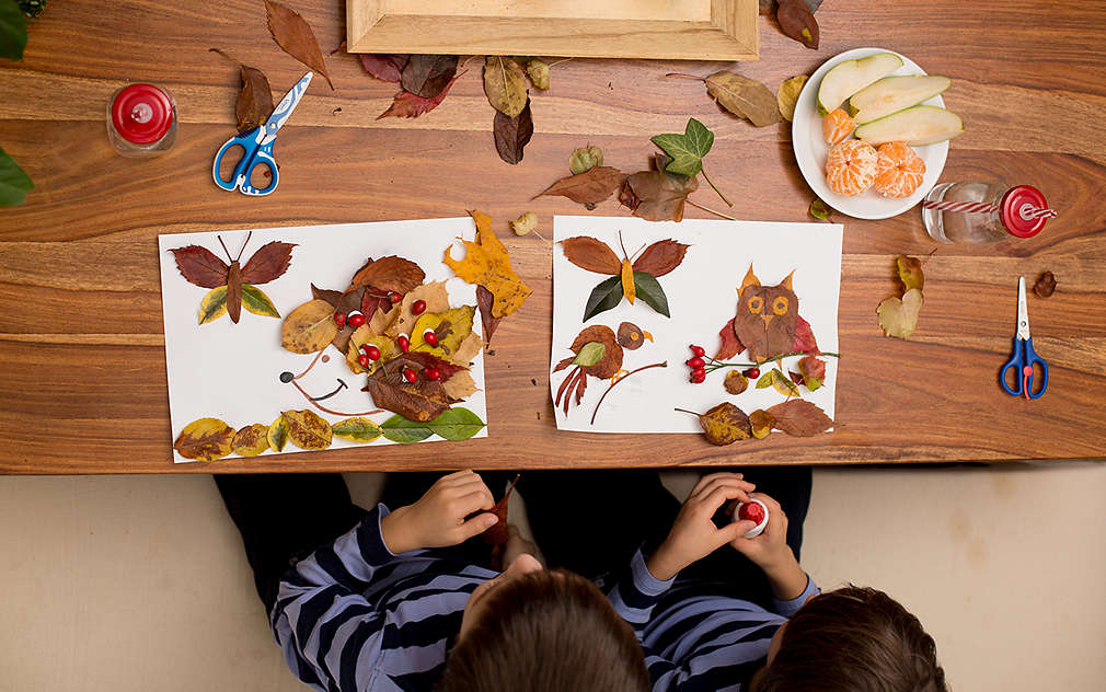 Prikazan je veliki smeđi drveni stol iz ptičje perspektive za kojim sjedi dvoje djece koja lijepe životinje od lišća na bijeli papir. Na stolu se nalaze pribor, lišće i tanjur s voćem te drvena zdjela s trima svjećicama.