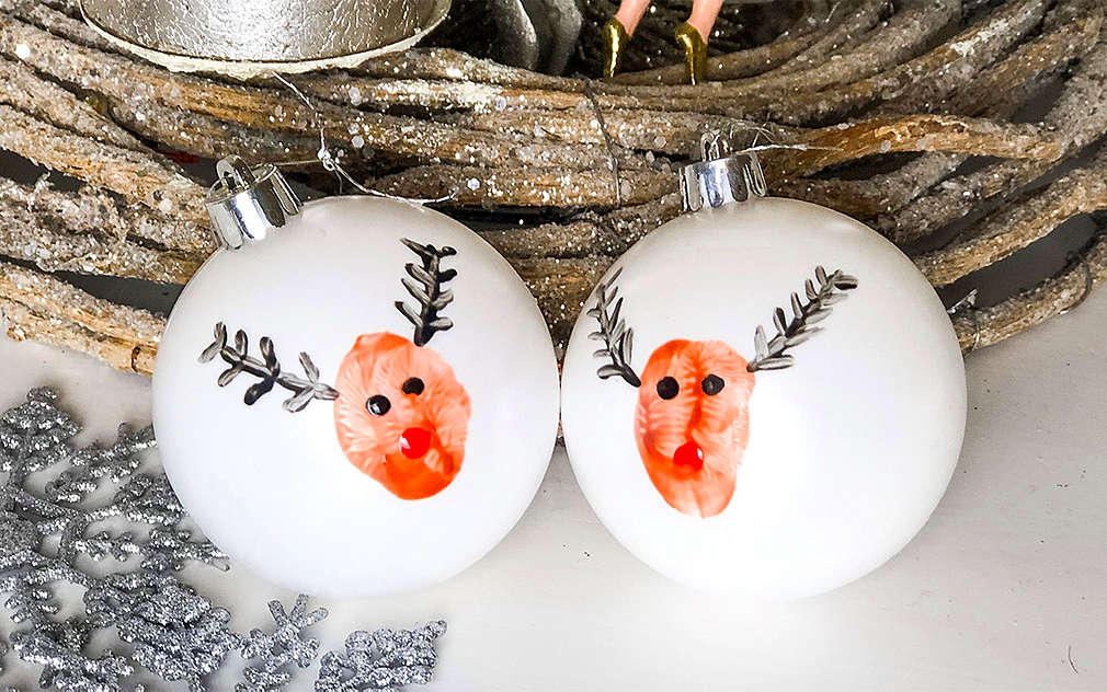 Dvě bílé vánoční koule, na kterých jsou vyobrazení vlastnoručně namalovaní sobi, v pozadí je věnec ze dřeva a stříbrná ozdoba na vánoční stromeček.