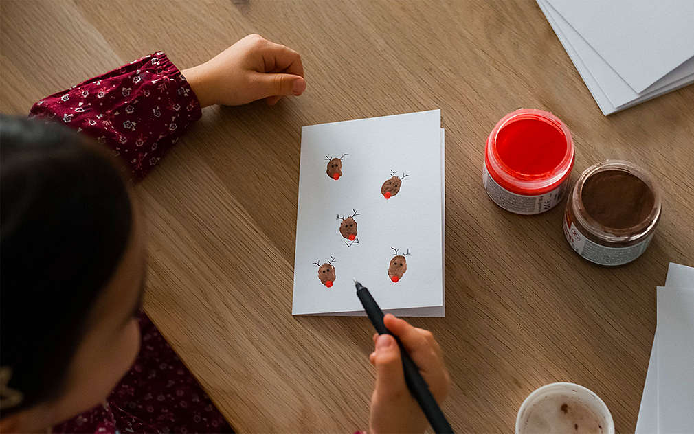 Malá holčička pomalovává bílé přání sobími motivy u dřevěného stolu, na kterém je další papír a červená a hnědá barva.