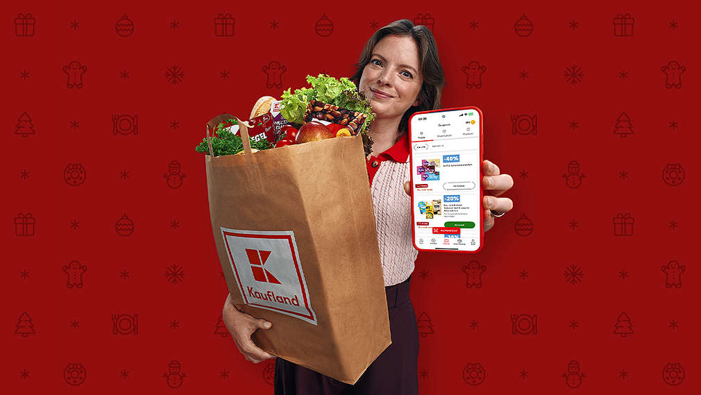 A woman holds a paper shopping bag with the Kaufland logo and a smartphone showing a coupon interface with a red Christmas-themed background.