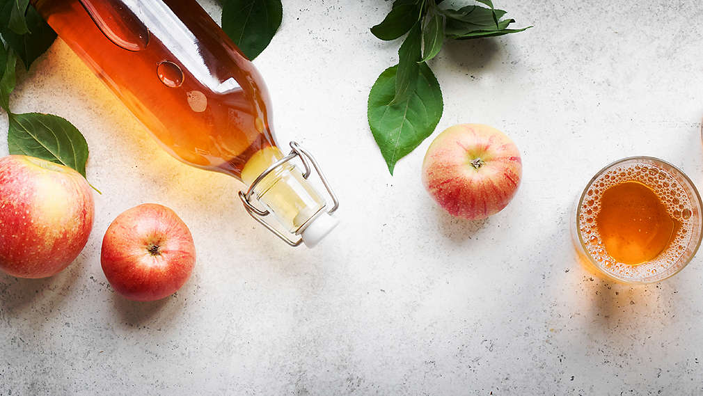 Изображена бутылка яблочного уксуса, яблоки и стакан с яблочным уксусом.