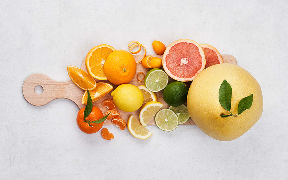 Na jasnoszarym blacie, na drewnianej desce leżą różne gatunki owoców cytrusowych, pokrojonych i w całości.
