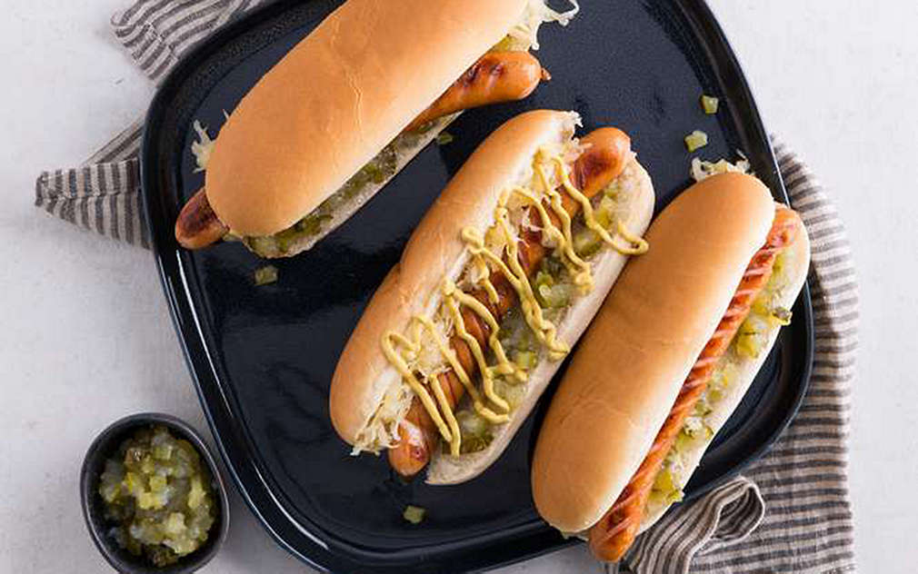 Na jasnym tle, na dużym czarnym talerzu leżą trzy hot dogi z frankfurterkami, relishem ogórkowym i musztardą. Obok stoi mała miseczka z relishem.