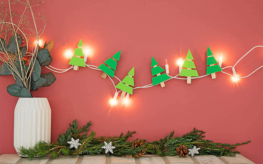Vianočné stromčeky vyrobené z penovej gumy a štipcov sú pripevnené na rozsvietenej LED svetelnej reťazi, ktorá je zavesená na ružovej stene.