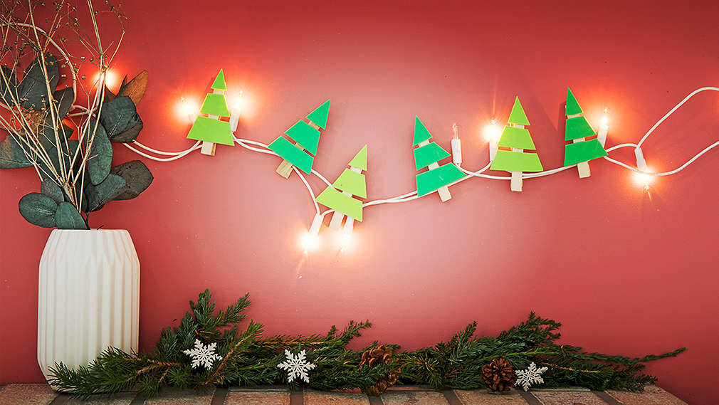 Na ružovej stene visí rozsvietená LED svetelná reťaz, ktorá je ozdobená vianočnými stromčekmi vyrobenými z mäkkej penovej gumy a štipcov. Naľavo je biela váza s vetvičkami.