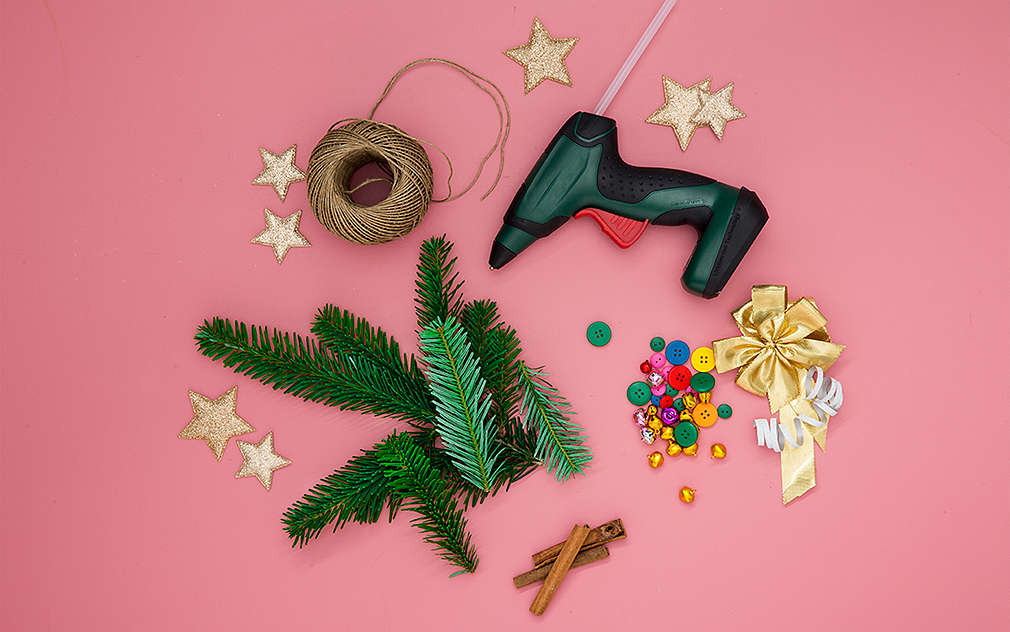 Na ružovom podklade sú položené jedľové vetvičky, tyčinky škorice, tavná lepiaca pištoľ a ďalšie pomôcky na tvorenie.