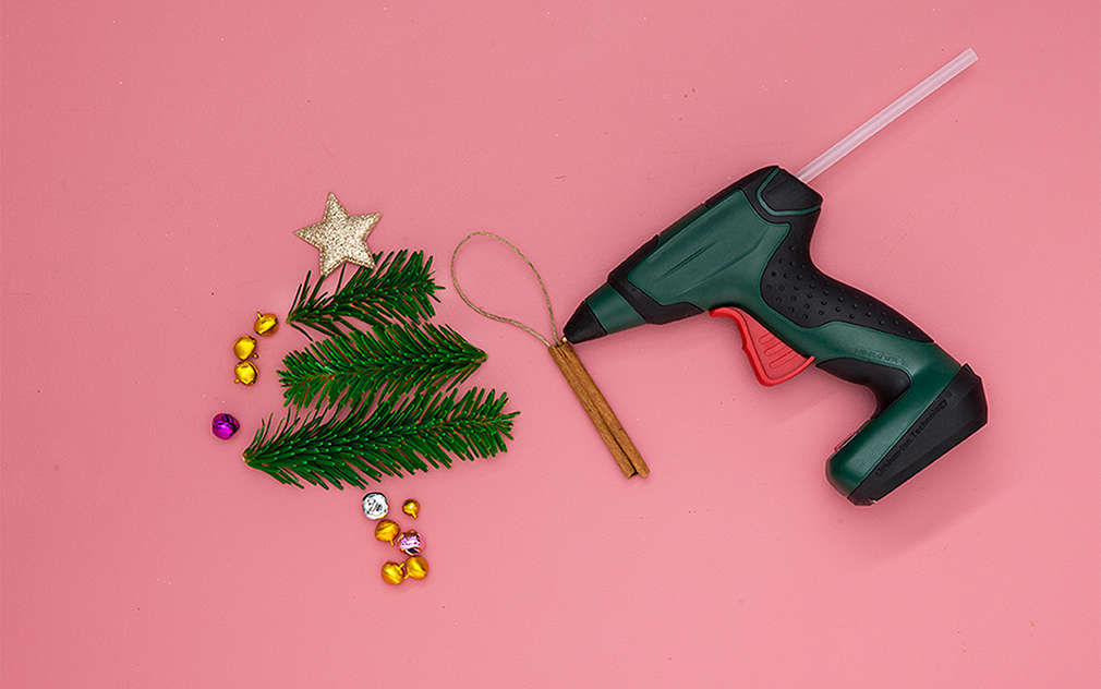 Na ružovom podklade je jedlička vyrobená z jedľových vetvičiek, jednej tyčinky škorice s očkom na zavesenie a ozdobných kameňov. Vedľa toho je položená tavná lepiaca pištoľ.