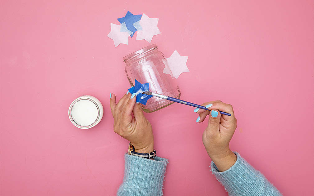 Dve ruky na ružovom podklade pripevňujú lepidlom modré a biele papierové hviezdy na priehľadný pohár.
