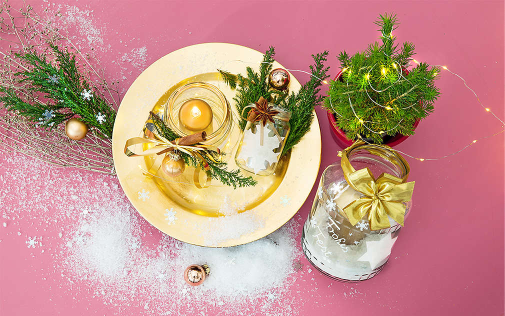 Vlastnoručne vyrobené vianočné svietniky sú položené na zlatom tanieri na ružovom podklade. Okolo sú rôzne vianočné ozdoby z jedľových vetvičiek, stromček a dekoratívny sneh.