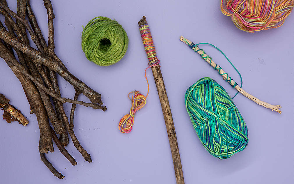 Drevená palička je omotaná farebnou vlnou a položená na fialovom podklade. Vedľa nej sú ďalšie klbká vlny a drevené paličky.