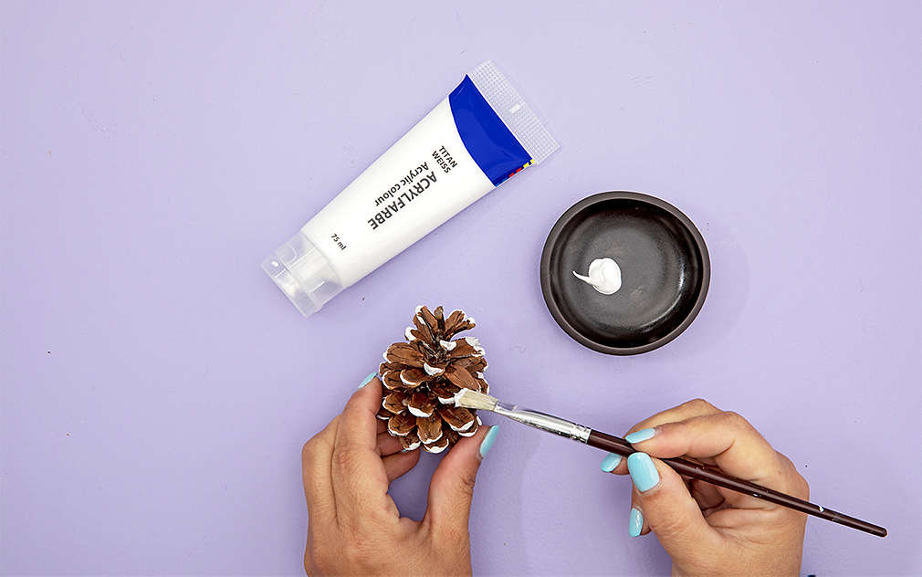 Dve ruky na fialovom podklade maľujú jedľovú šišku bielou farbou.