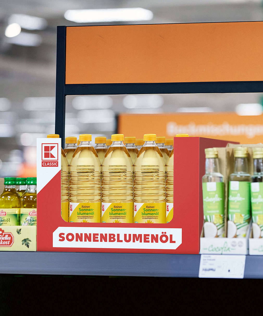 Изображение на регал в хипермаркет, на който има кашон с бутилки олио K-Classic