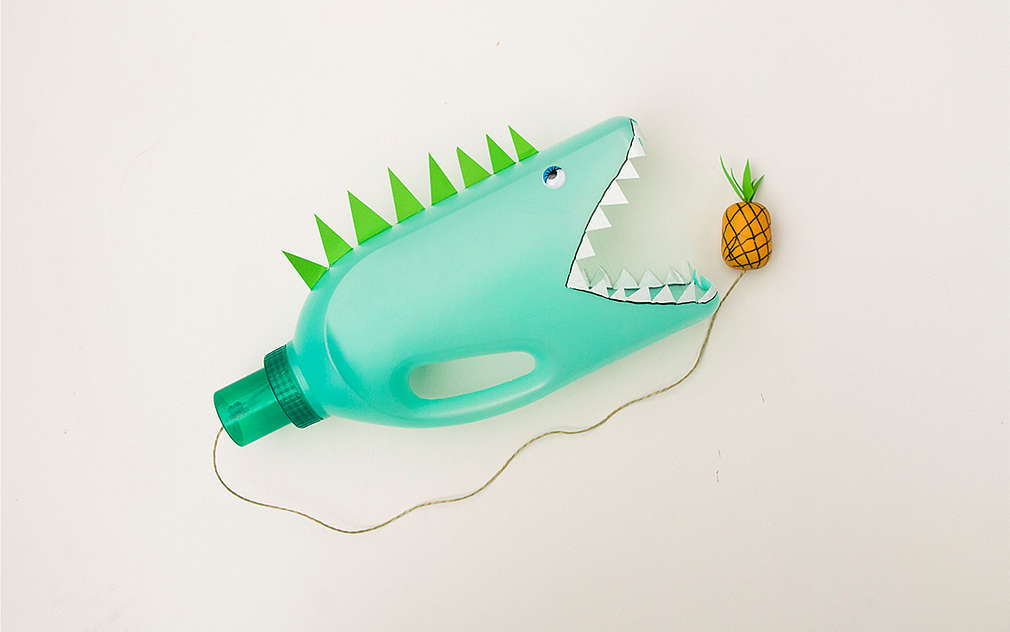 Jocul „aruncă și prinde mingea”, fotografiat de sus, arată ca un rechin și este realizat dintr-o sticlă de plastic verde. De sticlă atârnă o sfoară pe care se găsește un ananas.