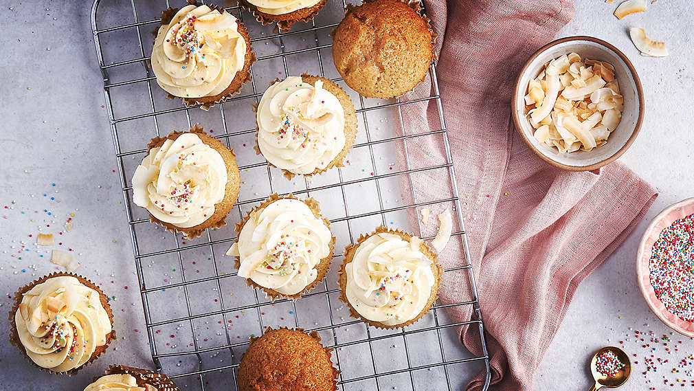 Viac ako desať cupcakes na mriežke na pečenie, pohľad zhora, na svetlosivom podklade. Väčšina má smotanovo biely krém. Okrem toho sa na ploche nachádza svetloružová kuchynská utierka, lyžica, miska so strúhaným kokosom a ďalšia miska s pestrofarebnými cukrovými guľôčkami.