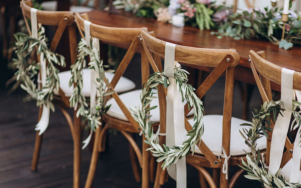 Masă de nuntă cu scaune din lemn cu perne de șezut albe, fiecare decorate cu o coroniță din ramuri, legate cu o fundă albă.