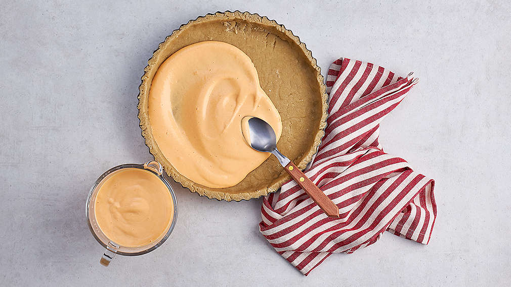 Teig in einer runden Kuchenform, darin wird eine cremige Füllung verteilt. Daneben eine Schüssel mit dieser Creme sowie ein weiß-rot gestreiftes Küchentuch., Teig in einer runden Kuchenform, darin wird eine cremige F�llung verteilt. Daneben eine Sch�ssel mit dieser Creme sowie ein wei�-rot gestreiftes K�chentuch.