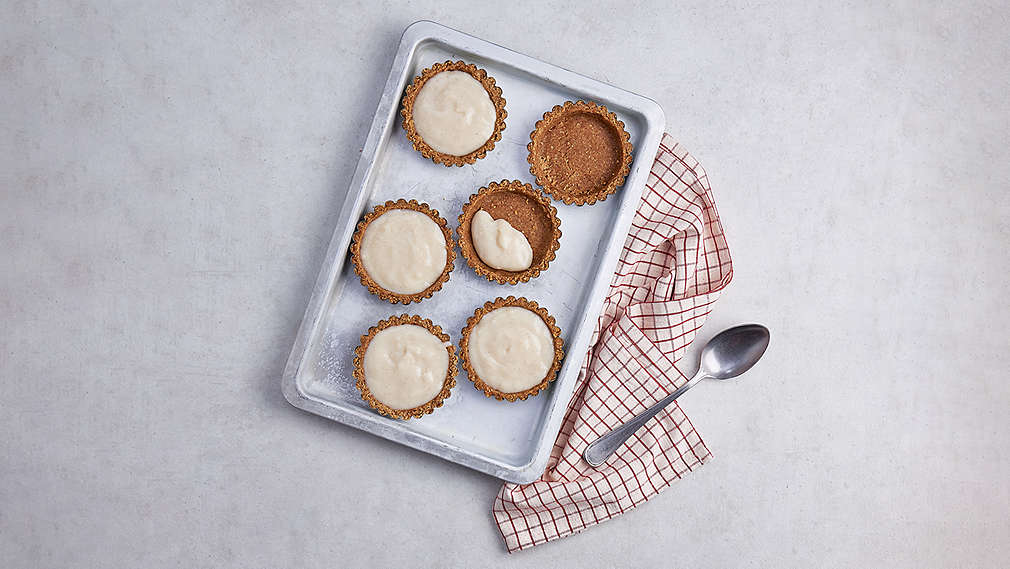 Kleine rude Teigformen auf einer Backform, gefüllt mit einer hellen Creme. Daneben ein karriertes Küchentuch sowie ein Löffel. , Kleine rude Teigformen auf einer Backform, gef�llt mit einer hellen Creme. Daneben ein karriertes K�chentuch sowie ein L�ffel.