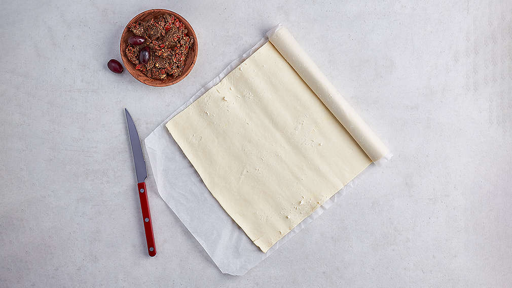 Ein zur Hälfte ausgerollter Blätterteig, daneben ein rotes Messer sowie eine Schüssel mit brauner Füllmasse., Ein zur H�lfte ausgerollter Bl�tterteig, daneben ein rotes Messer sowie eine Sch�ssel mit brauner F�llmasse.