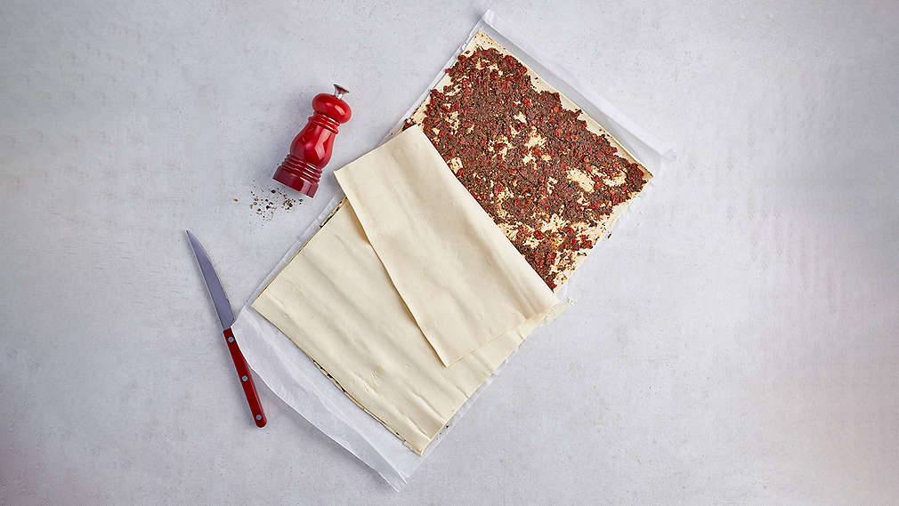 Mit brauner Füllmasse bestrichener Blätterteig. Daneben ein rotes Messer sowie eine rote Gewürzmühle. , Mit brauner F�llmasse bestrichener Bl�tterteig. Daneben ein rotes Messer sowie eine rote Gew�rzm�hle.