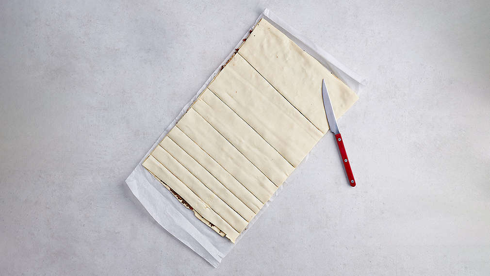 Vollständig zusammenkeklappter Blätterteig, welchen zum Teil in schmale Streifen geschnitten ist, daneben ein rotes Messer. , Vollst�ndig zusammenkeklappter Bl�tterteig, welchen zum Teil in schmale Streifen geschnitten ist, daneben ein rotes Messer.