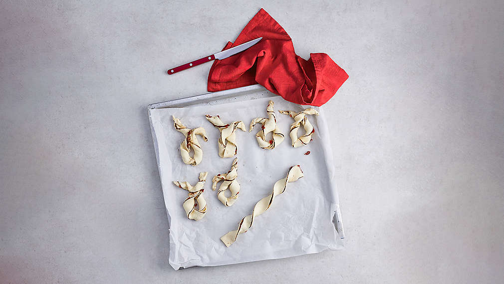 In Form von Hasen gedrehte Blätterteigstangen auf Backblech, daneben ein rotes Messer sowie ein rotes Küchentuch. , In Form von Hasen gedrehte Bl�tterteigstangen auf Backblech, daneben ein rotes Messer sowie ein rotes K�chentuch.