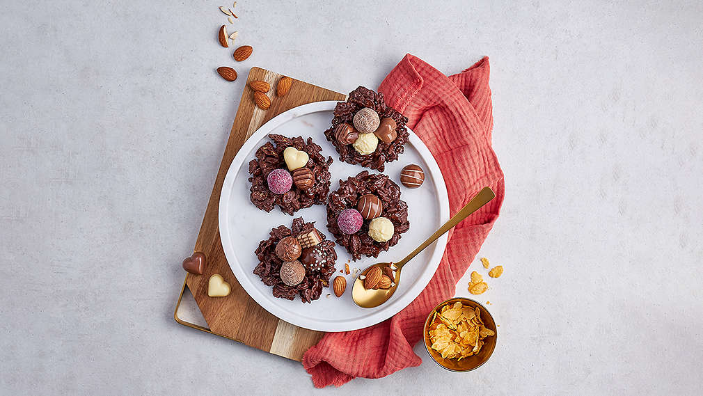 Schokoladennester, verziertmit bunten Pralinen auf einem weißen Teller. Daneben ein goldener Löffek, ein rotes Küchentuch sowie eine kleine Schüssel mit Cornflakes., Schokoladennester, verziertmit bunten Pralinen auf einem wei�en Teller. Daneben ein goldener L�ffek, ein rotes K�chentuch sowie eine kleine Sch�ssel mit Cornflakes.