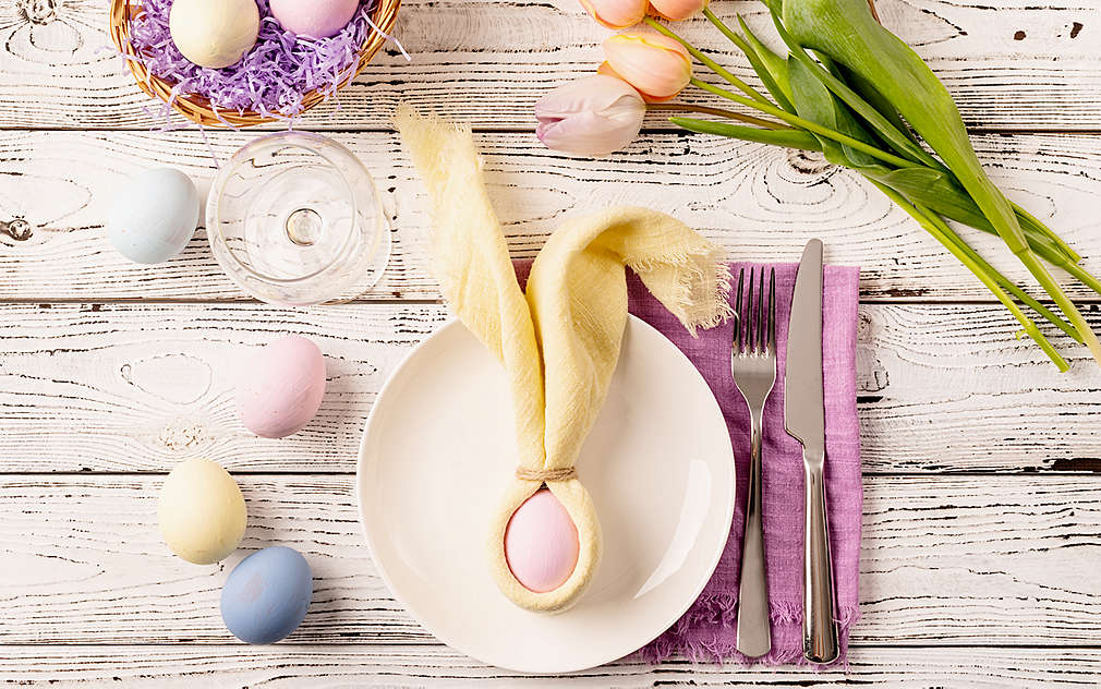 Na jasnym drewnianym stole znajduje się wiele wielkanocnych ozdób: pisanki malowane w pastelowych kolorach, tulipany oraz koszyczek wielkanocny z fioletową papierową trawą i dwiema malowanymi pisankami. Widok z góry na biały talerz, na którym leży płócienna serwetka złożona w uszy wielkanocnego zajączka w kolorze żółtym, którą owinięta jest bladoróżowa pisanka. Obok talerza stoi szklanka z wodą i leży fioletowa serwetka ze sztućcami.