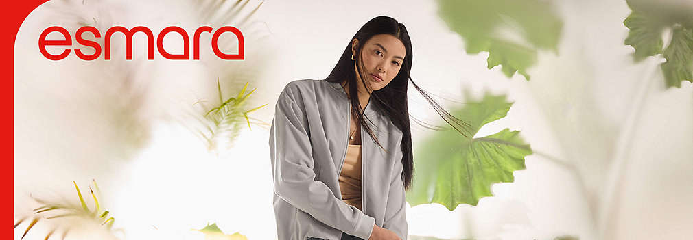 Eine junge Frau trägt eine hellgraue Jacke der Marke Esmara vor einer hellen Kulisse mit unscharfen, grünen Tropenpflanzen im Vordergrund., Eine junge Frau tr�gt eine hellgraue Jacke der Marke Esmara vor einer hellen Kulisse mit unscharfen, gr�nen Tropenpflanzen im Vordergrund.