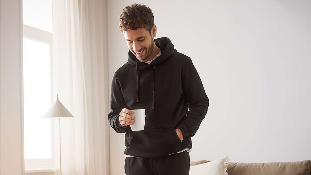 Ein lächelnder Mann mit lockigem Haar trägt einen gemütlichen schwarzen Kapuzenpullover und hält eine weiße Tasse in der Hand., Ein l�chelnder Mann mit lockigem Haar tr�gt einen gem�tlichen schwarzen Kapuzenpullover und h�lt eine wei�e Tasse in der Hand.