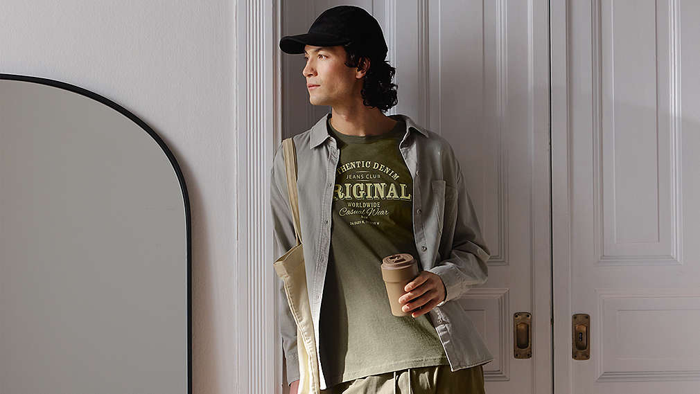 Ein junger Mann in einem olivgrünen Grafik-Shirt und hellem Hemd lehnt entspannt an einer Tür mit moderner Casual Wear, die nach Oeko-Tex Standards gefertigt wurde., Ein junger Mann in einem olivgr�nen Grafik-Shirt und hellem Hemd lehnt entspannt an einer T�r mit moderner Casual Wear, die nach Oeko-Tex Standards gefertigt wurde.