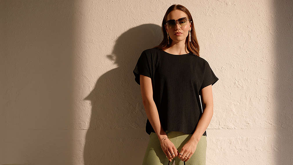 Eine Frau präsentiert moderne Esmara Umstandsmode, bestehend aus einem lockeren schwarzen T-Shirt und einer hellgrünen Hose, ergänzt durch eine Sonnenbrille und auffälligen Schmuck vor einer hellen Wand., Eine Frau pr�sentiert moderne Esmara Umstandsmode, bestehend aus einem lockeren schwarzen T-Shirt und einer hellgr�nen Hose, erg�nzt durch eine Sonnenbrille und auff�lligen Schmuck vor einer hellen Wand.