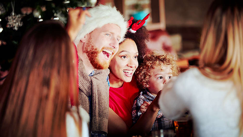 Un grup de oameni, în centru un băiat, o fată și un copil care sunt în spiritul Crăciunului. Băiatul poartă o căciulă de Moș Crăciun, fata poartă o bentiță de sezon, în fundal se vede un brad împodobit.