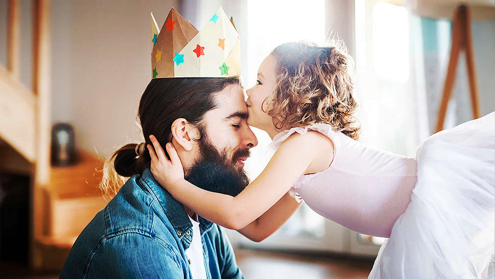 Un tată tânăr cu barbă și părul lung strâns poartă o coroană handmade și primește un sărut pe frunte de la fiica sa, care poartă o rochiță roz.