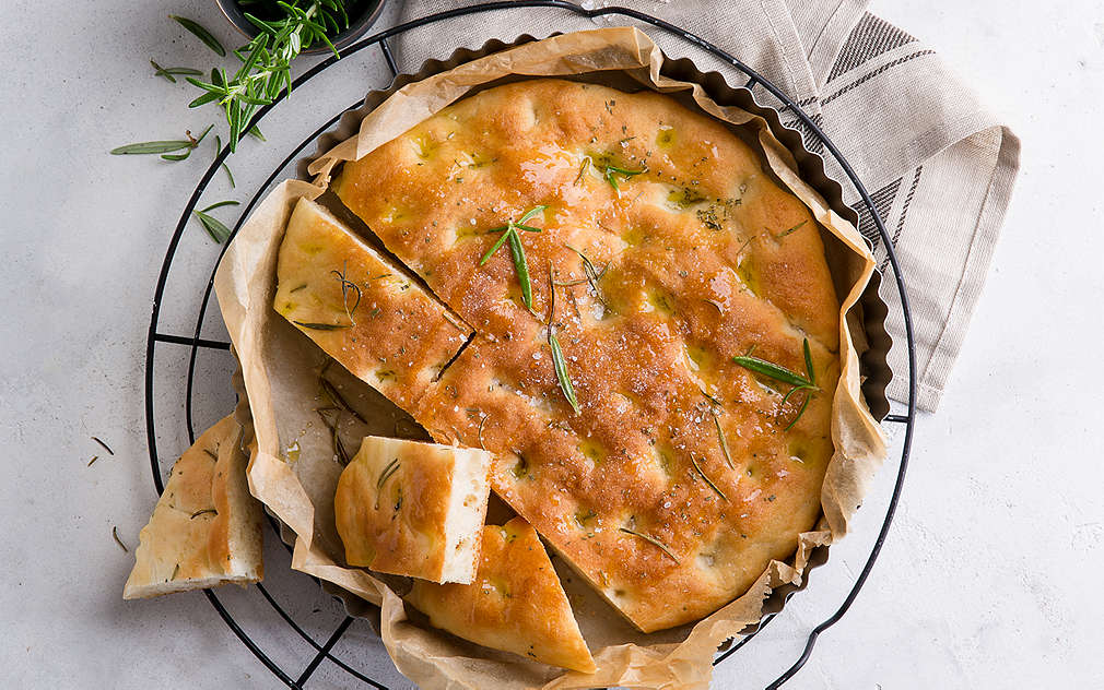 Na slici se vidi plosnata, okrugla focaccia na okrugloj rešetci ispod koje se nalazi bijelo-siva kuhinjska krpa. Lijevo od toga nalazi se kalup za pečenje iznad kojeg su dvije zdjelice, jedna s ružmarinom i jedna s krupnom soli.