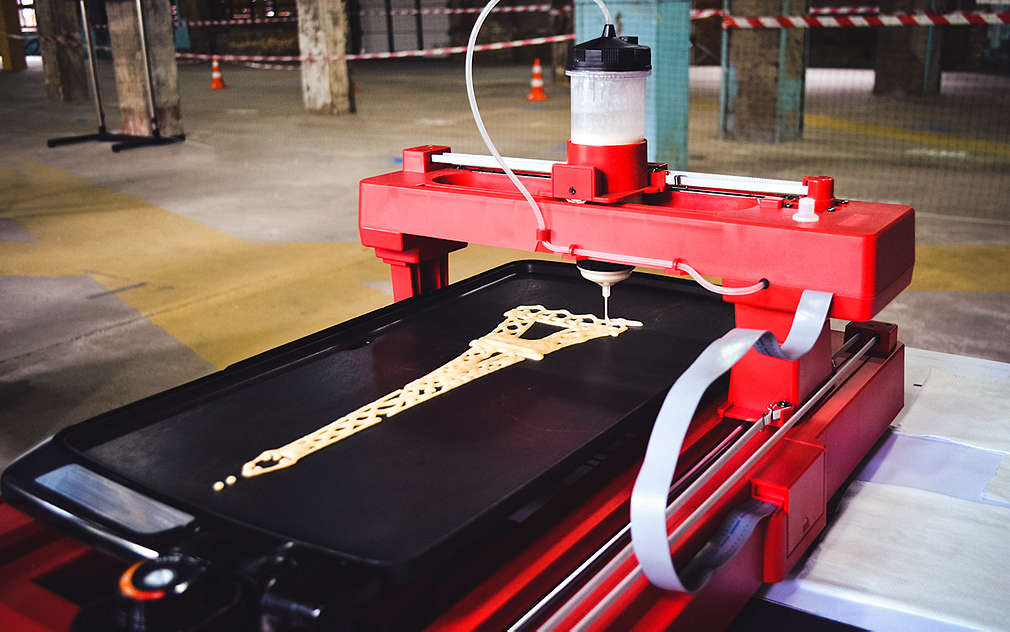 Duża drukarka spożywcza, przez której dyszę płynie ciasto i która robi naleśniki w kształcie wieży Eiffla na czarnej powierzchni.